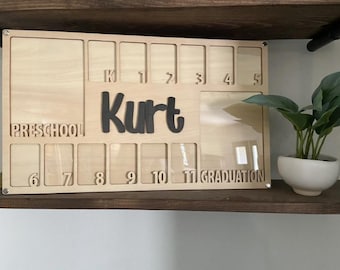May include: A wooden photo display board with 11 frames for pictures. The board is labeled with the numbers 1 through 11, with the first frame labeled "Preschool" and the last frame labeled "Graduation". The board also has the name "Kurt" written on it.
