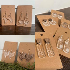 Op de afbeelding: Een verzameling acryl en houten oorbellen, gepresenteerd op bruine kaarten. De oorbellen hebben verschillende ontwerpen, waaronder boerderijdieren, bloempatronen en geometrische vormen. De oorbellen zijn zilverkleurig en hebben een heldere, transparante uitstraling.