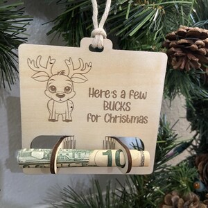 Może przedstawiać: Drewniana ozdoba choinkowa z ilustracją renifera i tekstem "Here's a few bucks for Christmas". W szczelinie ozdoby widoczny jest zwinięty banknot dolarowy.
