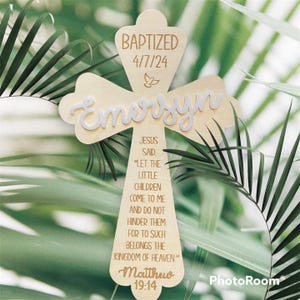 May include: A wooden cross with the name "Emersyn" in white script. The cross is inscribed with "BAPTIZED 4/7/24" and a Bible verse from Matthew 19:14. A small dove is etched above the date. The cross is set against a backdrop of green foliage.