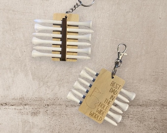 May include: Two golf tee keychains. One keychain has a wooden holder with white tees. The other keychain has a wooden tag with the text "BEST DAD IN THE HOLE WIDE WORLD" and a golf course graphic, with white tees.