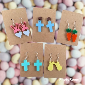 Puede incluir: Una colección de pendientes con temática de Pascua exhibidos en tarjetas marrones. Los pendientes presentan varios diseños, incluyendo conejos gnomos rosas, cruces azules, zanahorias naranjas y conejos amarillos. Los pendientes son de acrílico y tienen ganchos plateados.