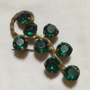 May include: Vintage brooch featuring seven round, emerald green gemstones set in a gold-toned metal vine design. The brooch has a pin closure on the back. A classic piece of jewelry.