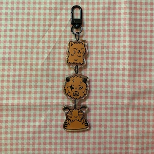 May include: A keychain featuring three orange cartoon character charms. The charms are connected by small black rings and a black clasp. The characters have a variety of expressions and are outlined in black.