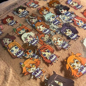 May include: A collection of anime-style character stickers. Each sticker features a chibi-style girl with large eyes, wearing a plaid dress. The stickers have various hair colors, including orange, brown, and white. The background is a light pink.