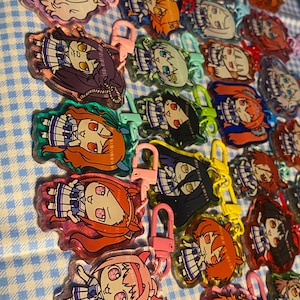 May include: A collection of colorful acrylic keychains featuring anime-style character designs. Each keychain has a different character with varying hair colors and outfits, attached to a metal clasp. The keychains are arranged on a blue and white checkered surface.