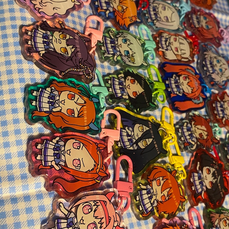 May include: A collection of colorful acrylic keychains featuring anime-style character designs. Each keychain has a different character with varying hair colors and outfits, attached to a metal clasp. The keychains are arranged on a blue and white checkered surface.