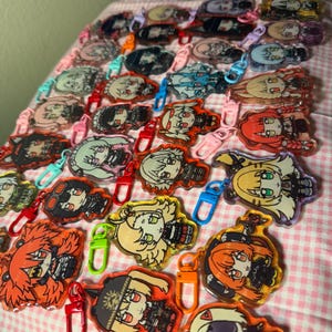 May include: A collection of colorful acrylic keychains featuring anime-style character designs. Each keychain has a unique character illustration with vibrant colors and attached to a metal clasp. The keychains are arranged on a pink and white checkered surface.