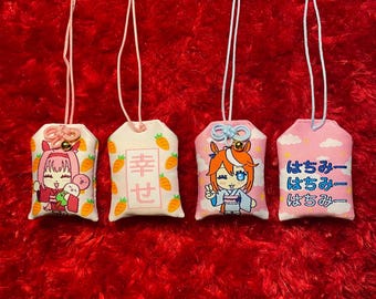 May include: Four Japanese omamori charms with white cords hang against a red background. The charms are rectangular with rounded tops. Each charm has a different design, including cartoon characters, carrots, and Japanese text.