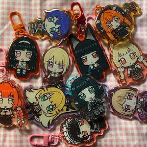 May include: A collection of colorful anime-style character keychains. Each keychain features a unique character design with vibrant colors, attached to a metal clasp. The keychains are arranged on a pink and white checkered surface.