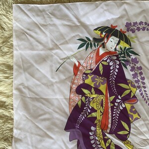 May include: A white fabric square with a colorful illustration of a woman in a traditional Japanese kimono holding a branch of wisteria flowers. The woman is wearing a purple and yellow kimono with a red sash. The wisteria flowers are purple and white.
