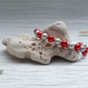Peut inclure: Un bracelet de perles alternant des perles rouges et argentées repose sur une roche claire et texturée. Le bracelet est un simple fil de perles rondes, sur un fond flou de couleur claire.