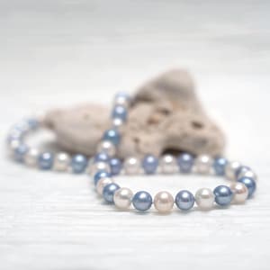 May include: A strand of white and blue pearls on a white background. The pearls are arranged in a circle and are strung on a thin wire.