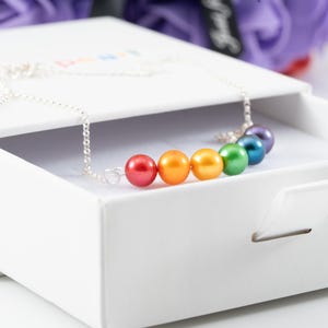 May include: A silver chain necklace featuring a row of six round, smooth beads in a rainbow gradient of red, orange, yellow, green, blue, and purple. The necklace is displayed resting on the edge of a white, rectangular, sliding jewelry gift box.