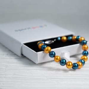Puede incluir: Un brazalete hecho de perlas doradas y azules alternas en una caja blanca.