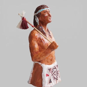 Puede incluir: Figurilla artesanal de un nativo americano, que representa una figura con una expresión serena, que lleva una diadema y vestimenta tradicional. La figura sostiene un bastón ceremonial con detalles en rojo, blanco y beige. El tono de piel de la figura es marrón cálido.