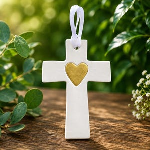 May include: A white ceramic cross ornament featuring a centered gold heart detail. The cross has a smooth, matte finish and includes a white ribbon loop at the top for hanging. The design is simple, with clean lines and a balanced, symmetrical shape.