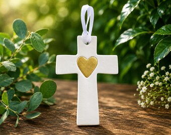 May include: A white ceramic cross ornament featuring a centered gold heart detail. The cross has a smooth, matte finish and includes a white ribbon loop at the top for hanging. The design is simple, with clean lines and a balanced, symmetrical shape.