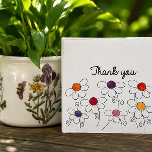 May include: A square white greeting card with the text Thank you in black script, featuring a hand-drawn illustration of several daisies with colorful centers in orange, purple, red, pink, and yellow, positioned next to a white ceramic plant pot.