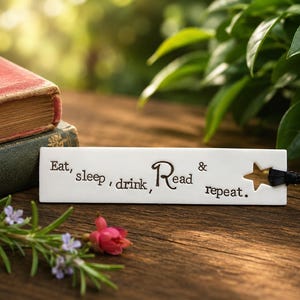 May include: A rectangular white ceramic bookmark featuring the black engraved text Eat, sleep, drink, Read & repeat. The bookmark has a star-shaped cutout on the right side and a thin black ribbon attached through the opening.