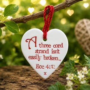 Op de afbeelding: Een wit hartvormig keramisch ornament dat aan een rood koord hangt. De voorzijde is voorzien van rode reliëftekst met de tekst: A three cord strand isn't easily broken. Ecc 4:12 x. Het ornament heeft een gladde, glanzende afwerking.