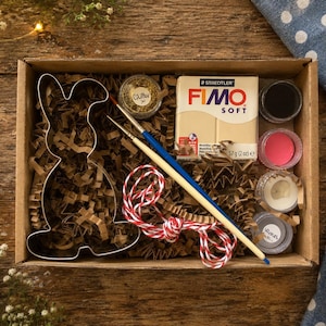 May include: A craft kit in a cardboard box containing a metal rabbit-shaped cookie cutter, a block of beige Staedtler Fimo Soft modelling clay, two paintbrushes, a small container of gold glitter, four small paint pots, and red and white twine.