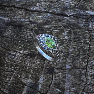 May include: A silver ring featuring a green gemstone surrounded by smaller blue gemstones. The ring is resting on a wooden surface.