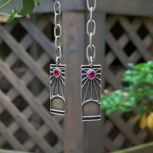 May include: Silver dangle earrings with a pink gemstone and a sunburst design. The earrings are hanging from a chain.