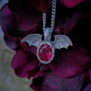 Könnte beinhalten: Eine silberne Fledermaus-Anhängerkette mit einem großen roten Edelstein. Der Anhänger hängt an einer silbernen Kette.