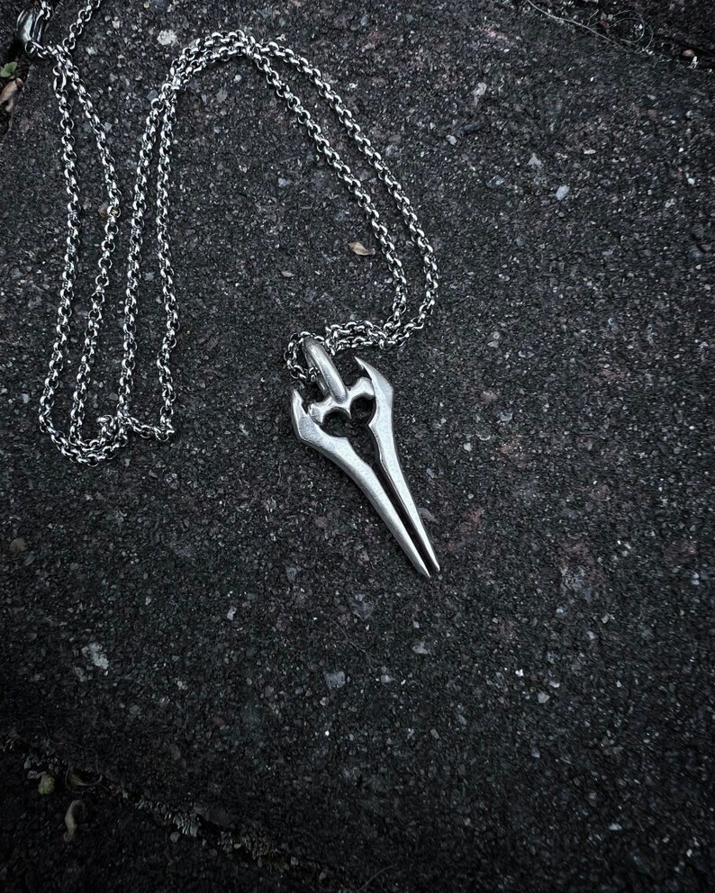Puede incluir: Un collar de plata con un colgante con forma de espada. El colgante tiene un dise&ntilde;o detallado con una punta afilada y un mango curvo.