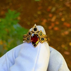 Puede incluir: Un anillo de oro con una piedra roja en forma de pera rodeada de piedras negras m&aacute;s peque&ntilde;as. El anillo est&aacute; engastado en un dise&ntilde;o geom&eacute;trico.