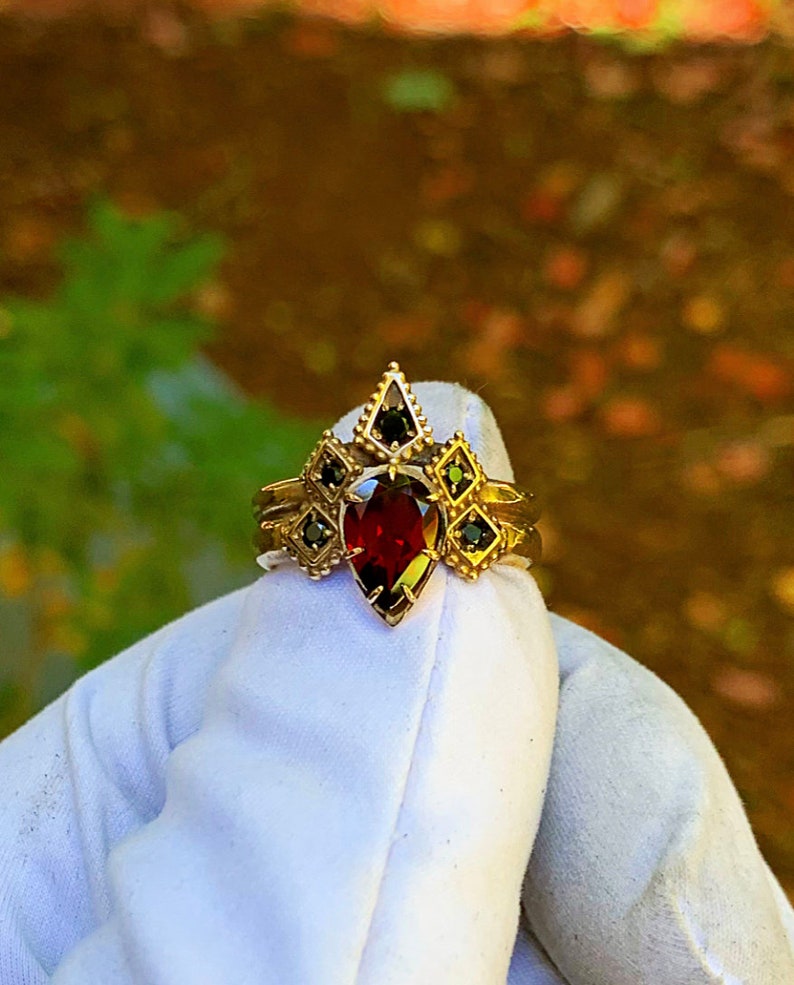 Puede incluir: Un anillo de oro con una piedra roja en forma de pera rodeada de piedras negras m&aacute;s peque&ntilde;as. El anillo est&aacute; engastado en un dise&ntilde;o geom&eacute;trico.