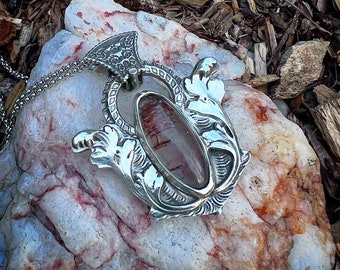 Puede incluir: Un colgante de plata con un diseño ornamentado que presenta una piedra de cuarzo transparente con inclusiones rojas. El colgante está en una cadena de plata y descansa sobre una roca con un patrón de mármol rosa y blanco.