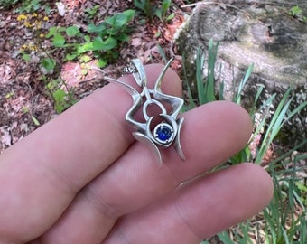 Puede incluir: Un colgante plateado con un diseño abstracto, con una gema azul central. El colgante se sostiene en una mano, con un anillo negro visible en un dedo. El fondo muestra vegetación y un tocón de árbol.