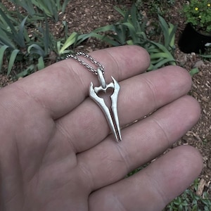 Puede incluir: Un colgante de color plateado con forma de espada de doble filo. El colgante est&aacute; en una cadena de plata.