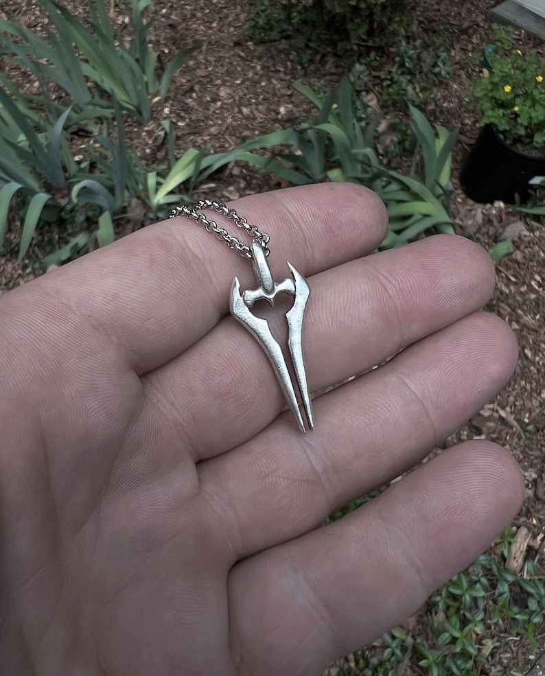 Puede incluir: Un colgante de color plateado con forma de espada de doble filo. El colgante est&aacute; en una cadena de plata.