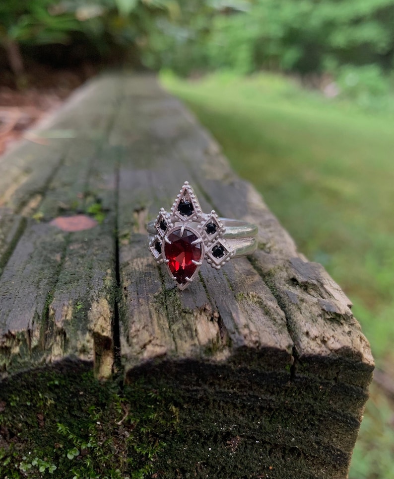 Puede incluir: Un anillo de plata con una piedra preciosa roja en forma de pera rodeada de piedras negras m&aacute;s peque&ntilde;as. El anillo est&aacute; sobre una superficie de madera.