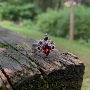 Puede incluir: Un anillo de plata con una piedra roja en forma de l&aacute;grima rodeada de piedras negras m&aacute;s peque&ntilde;as. El anillo est&aacute; sobre una superficie de madera.