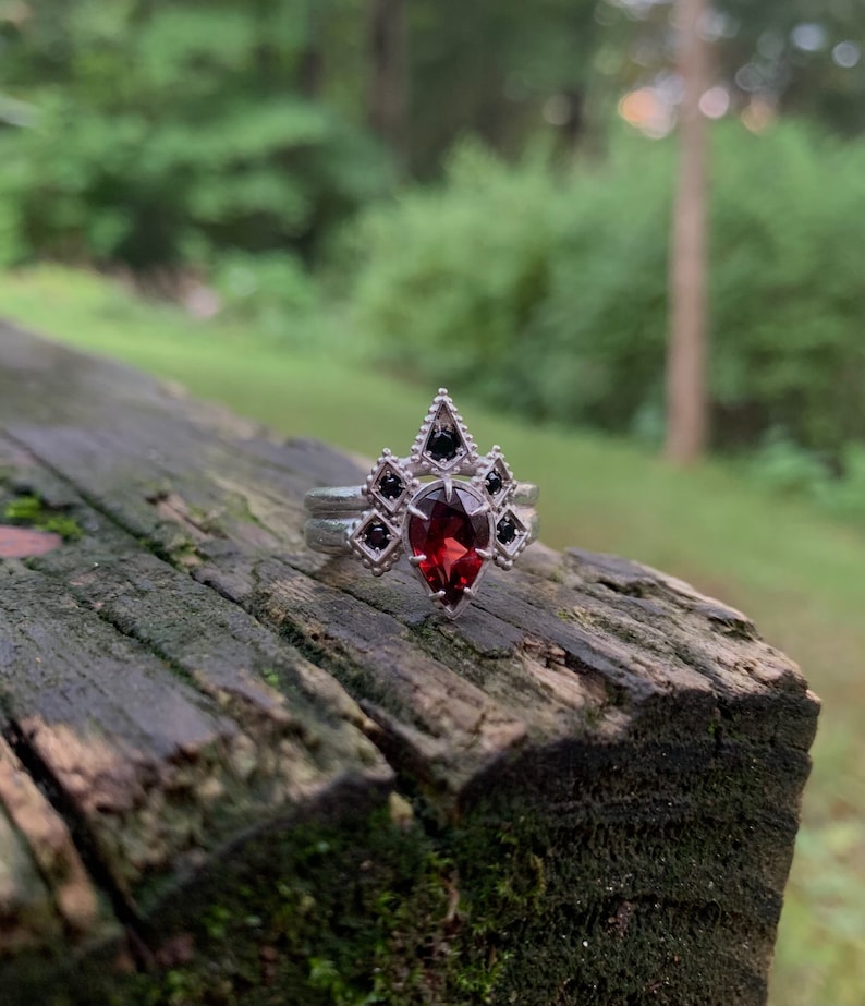 Puede incluir: Un anillo de plata con una piedra roja en forma de l&aacute;grima rodeada de piedras negras m&aacute;s peque&ntilde;as. El anillo est&aacute; sobre una superficie de madera.