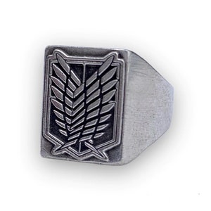 Può includere: Un anello d'argento con un emblema nero e argento di un simbolo alato. L'anello è di forma quadrata con bordi arrotondati.
