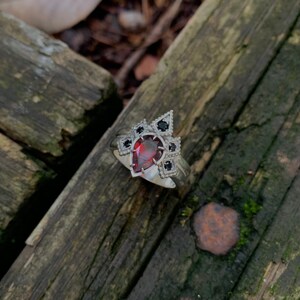 Puede incluir: Un anillo de plata con una piedra preciosa roja en forma de pera rodeada de piedras preciosas negras en un dise&ntilde;o geom&eacute;trico.