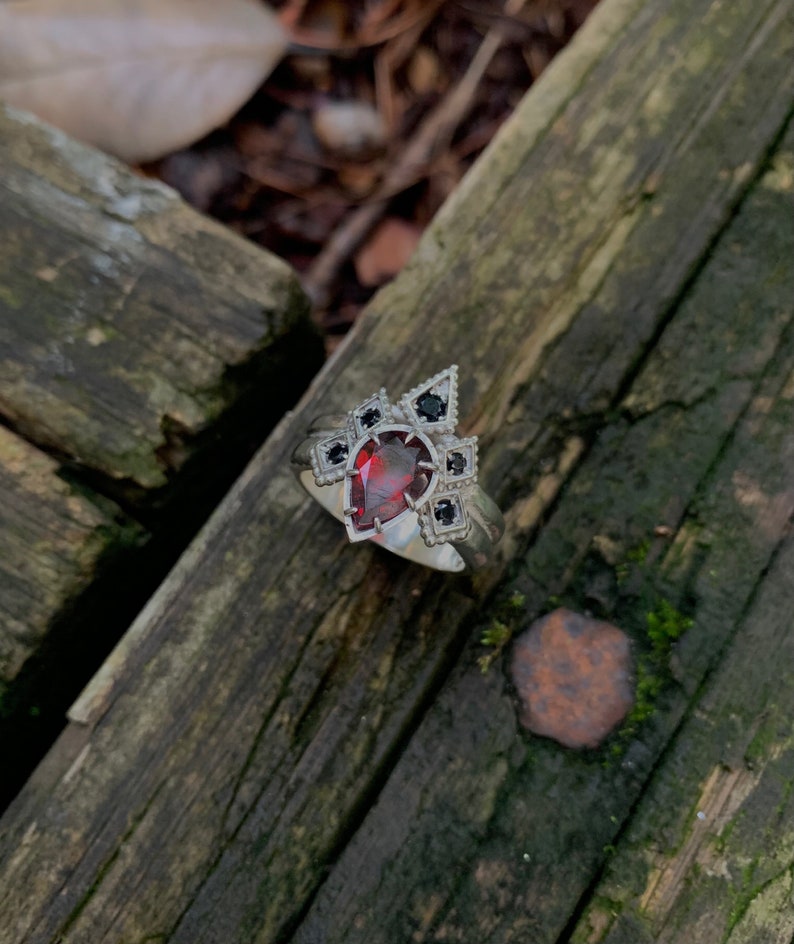Puede incluir: Un anillo de plata con una piedra preciosa roja en forma de pera rodeada de piedras preciosas negras en un dise&ntilde;o geom&eacute;trico.
