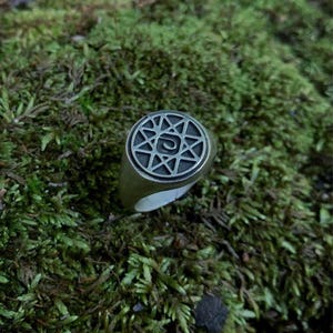 Puede incluir: Un anillo plateado con un diseño circular que presenta un patrón en forma de estrella. El anillo se encuentra sobre un fondo de musgo verde, creando una estética natural y terrosa. El diseño del anillo es detallado e intrincado.
