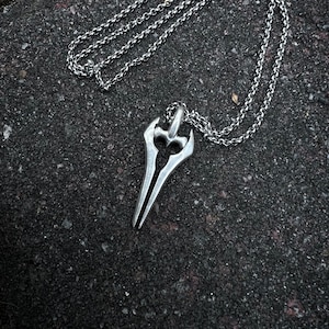 Puede incluir: Un collar de cadena de plata con un colgante con forma de espada. El colgante tiene una punta afilada y un mango curvo.