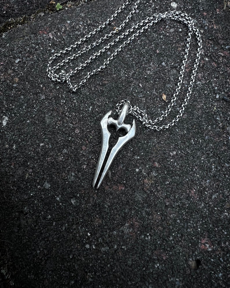 Puede incluir: Un collar de cadena de plata con un colgante con forma de espada. El colgante tiene una punta afilada y un mango curvo.
