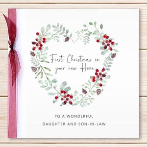 May include: A square greeting card with a white background featuring a watercolor heart-shaped wreath of green leaves, red berries, and small pinecones. The text reads First Christmas in your new Home and TO A WONDERFUL DAUGHTER AND SON-IN-LAW. A sheer dark red ribbon is tied on the left side.