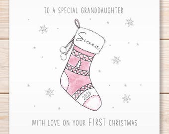 May include: A square greeting card featuring a pink Christmas stocking illustration with the name Sienna, the year 2026, and a small tree icon. The text reads TO A SPECIAL GRANDDAUGHTER and WITH LOVE ON YOUR FIRST CHRISTMAS.