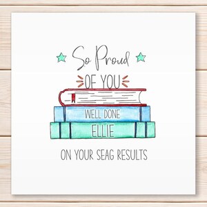 May include: A white card featuring a watercolour illustration of three books, stacked. The top book is red, the middle is blue, and the bottom is green. Text reads "So Proud of You" and "Well Done Ellie On Your SEAG Results".