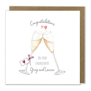 May include: A white card with a hand-drawn illustration of two champagne flutes clinking together. The card says "Congratulations on your engagement Greg and Lauren" with two small pink hearts above the flutes.
