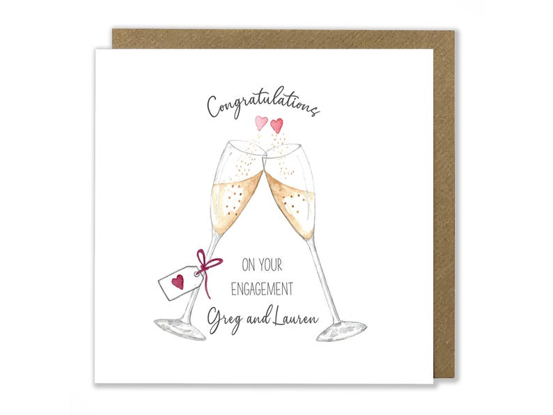 May include: A white card with a hand-drawn illustration of two champagne flutes clinking together. The card says "Congratulations on your engagement Greg and Lauren" with two small pink hearts above the flutes.